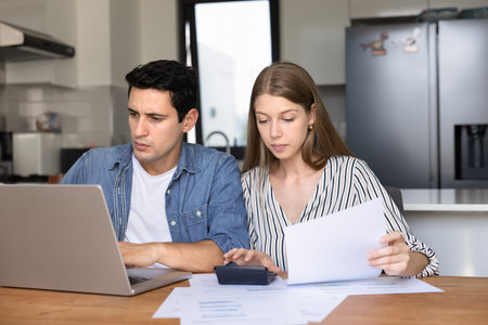 Young couple sits in modern kitchen reviewing financial documents, using laptop and calculator,...