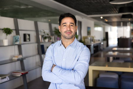 Confident handsome young professional man posing in office workspace, standing in co-working, keeling arms crossed in gesture of success, looking at camera, posing for head shot portraitの写真素材