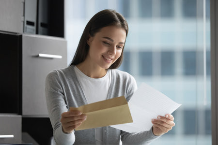 Reading good business news. Happy smiling young businesswoman company manager pull paper letter out of postal envelope enjoy successful job offer project approval promotion at stylish modern officeの写真素材