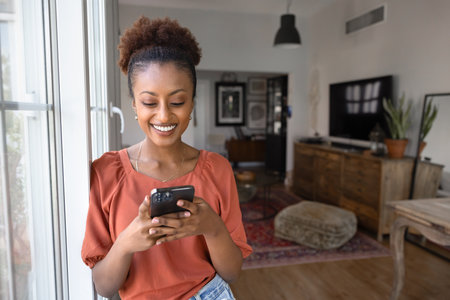 Positive beautiful young Black woman using online app on mobile phone, standing at window in cozy stylish home apartment, enjoying communication, chatting with toothy smile, browsing Internetの写真素材