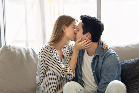 Young romantic couple in casual clothes sit close together on cozy couch, kissing, enjoying moment of closeness, reflecting love, warmth, and deep emotional connection. Dating, relationships, passionの写真素材