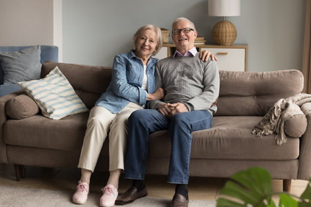 Family happiness and harmony. Full length portrait happy elderly married couple sit close together on sofa. Smiling mature wife hug shoulders of hoary husband looking at camera with affectionate smileの写真素材