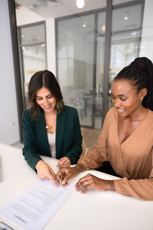 Positive confident young Hispanic business woman giving contract to African partner for signing, pointing finger at space for signature, starting collaboration, partnership. Vertical shotの写真素材