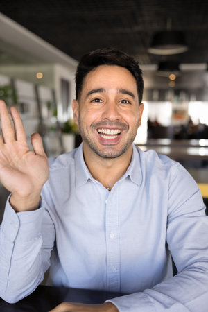 Happy young Latin businessman saying hi on video call, waving greeting hand hello at camera, talking...