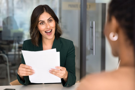 Excited positive HR manager woman meeting with job applicant at office table, holding paper documents, resume, learning about spectacular professional skills of candidate, gasping in surpriseの写真素材