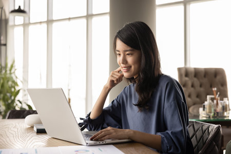 Positive young Asian analyst woman working at computer in co-working alone, typing on laptop,...