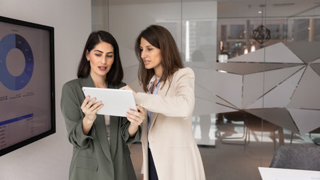 Two professional women collaborating in office, analyze data on digital tablet, engaged in...
