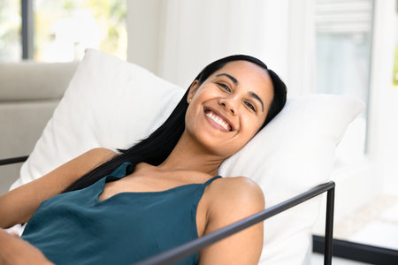 Happy young indigenous woman resting on soft comfortable trendy couch, looking at camera with toothy smile, enjoying relaxation, leisure, break, posing for homey portraitの写真素材