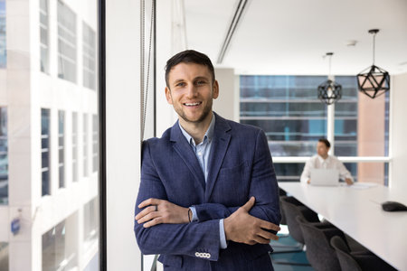 Positive Caucasian businessman dressed in formal suit stands in modern office meeting room, looking at camera with joyful expression, feel satisfied with career, exudes competence and professionalismの写真素材