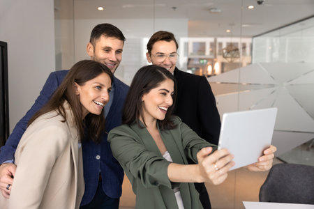Four happy multiethnic colleagues gather closely to take cheerful group selfie on digital tablet,...