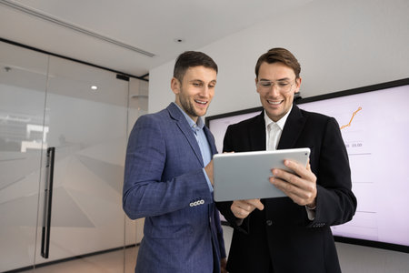 Two businessmen standing in modern office, smiling while review positive result on tablet, exuding satisfaction with sales growth, discussing successful project data, business progress and achievementの写真素材