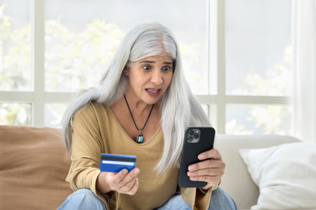 Mature woman holds card and smartphone, feels concerned, experiencing technical issues, unexpected...