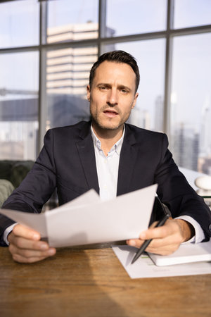 Confident serious Hispanic business leader man discussing legal or financial documents on work online conference chat, holding papers at workplace, speaking from screen. Vertical video call head shotの写真素材