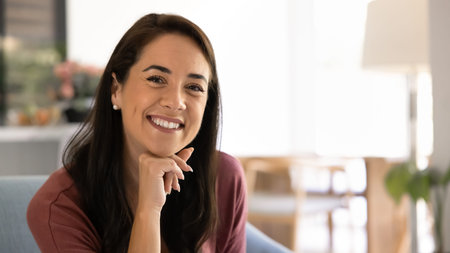 Close up portrait of cheerful middle-aged 40s brunette woman sits alone on cozy sofa at home,...