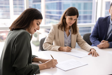 Two professional women signing contracts during formal business meeting seated at modern office table in conference room. Mutual agreement, commitment, trust between business parties, corporate dealの写真素材