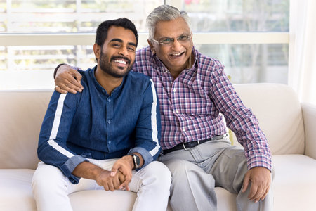 Older grandpa hugging his young adult Indian grandson, sit together on sofa, laughing, looking away....