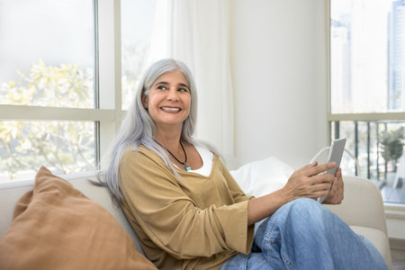 Happy middle-aged woman with long, silver hair sit comfortably on couch in cozy living room, holding...
