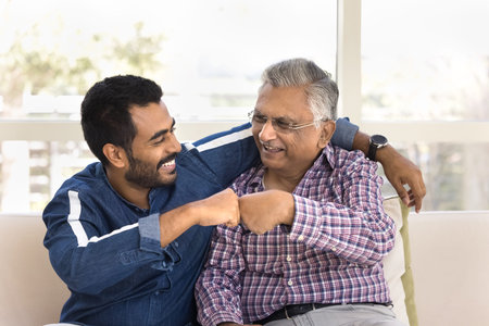 Happy young Indian man hugging and fist bumping older grandpa or great-grandfather seated together on couch, showing unity, understanding, sharing support. Intergenerational family ties, mutual respectの写真素材