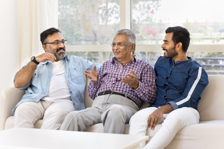 Smiling elderly Indian man sharing life story to middle-aged son and grown-up grandson sit in sofa...
