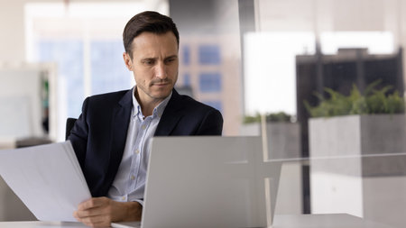 Focused man sits at desk, looks at laptop screen while holding sheet of paper, engaged in task...