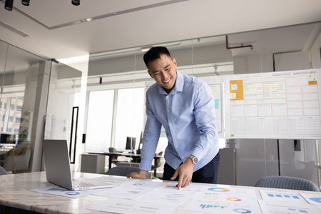 Smiling Asian professional man stands in modern office, looks at several papers spread across table,...