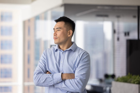 Serous Chinese professional man dressed in shirt standing alone in contemporary office with...