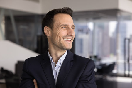Head shot of middle-aged businessman posing in modern workplace, looks into distance with joyful and contented expression, enjoys career growth, feel proud and satisfied with professional achievementsの写真素材