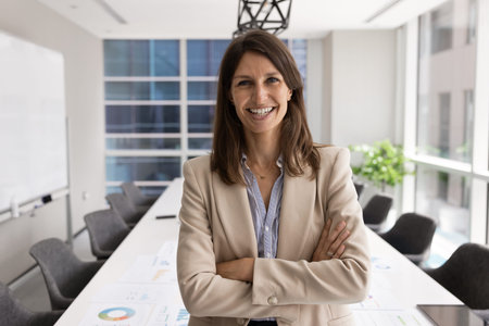 Middle-aged 40s confident businesswoman standing in modern conference room with her arms crossed,...