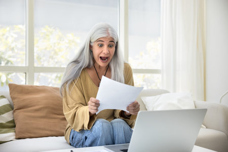 Mature woman gawp with mount opened at letter, reading document with surprised or shocked expression, reacting to unexpected information, review financial documents, work results, or personal mattersの写真素材