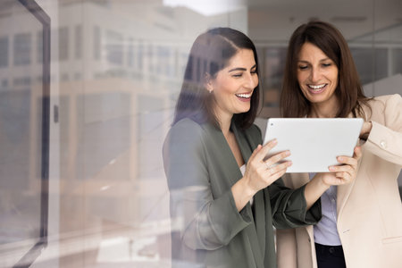 Two positive businesswomen using digital tablet together in office, smile while sharing creative ideas, reviewing amusing information, watch funny digital content during break using modern technologyの写真素材