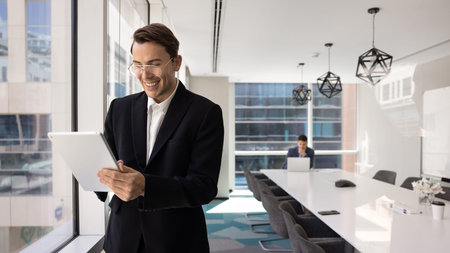 Confident young 35s businessman in suit smiling while working on digital tablet, reviewing...