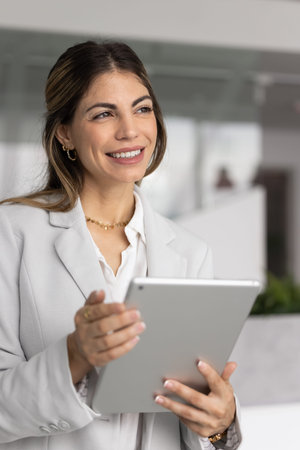Smiling young businesswoman standing in modern office with tablet looking away visualizing next steps in digital work creating new idea planning startup imagine career growth. Vertical photoの写真素材