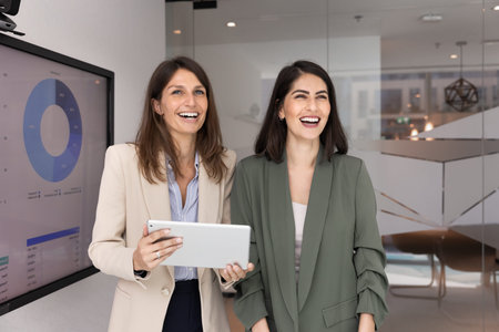 Two laughing women teammates standing in corporate office with tablet next to large digital screen...
