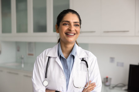 Confident young 30s Indian woman doctor dressed in white coat posing in clinic office, smiling,...