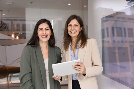 Portrait of two confident multiethnic businesswomen stand together in modern office, smiling at...