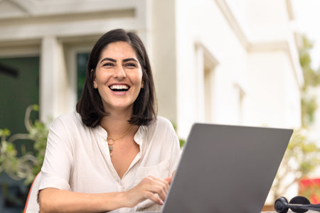 Cheerful young Hispanic remote worker woman sitting at workplace in yard outside of house, looking...