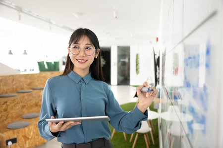Positive young Asian businesswoman in glasses working on project plan at Kanban board, writing tasks...