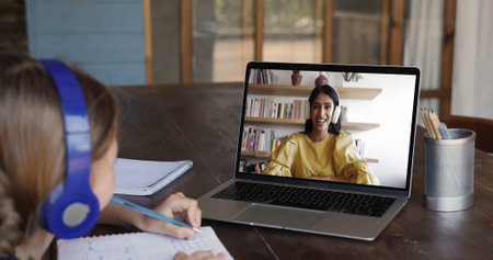 Kid girl wear headphones sit at desk, write in notebook while engaged in online lesson led by...