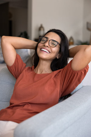 Young 30s woman leaning back in armchair with smile, hands resting behind her head in carefree,...