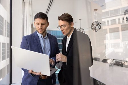 Two businessmen stand in modern conference room, deeply focused on laptop screen, colleague explain...