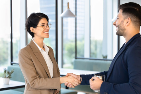 Two smiling multiethnic coworkers greeting during work break in office, shaking hands, starting...