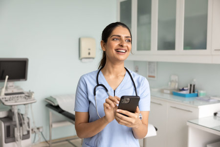 Smiling young female Indian nurse or GP doctor in blue scrubs with stethoscope holding smart phone in modern clinic exam room. Telehealth, patient support, medical communication, digital healthcareの写真素材