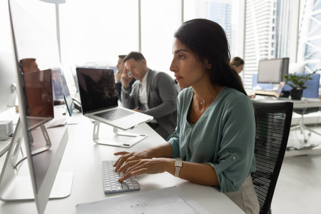 Focused Latina woman company department staff member typing on desktop pc working on report...