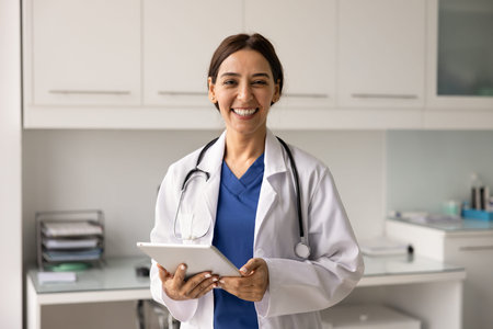 Portrait smiling female cardiologist doctor holds tablet, stands in modern clinic office, looks at...