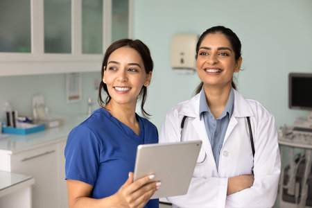 Two young attractive Indian and Arabian women doctors in uniforms standing in examination room with tablet, look into distance with contented, optimistic smile. Mission, healthcare services, teamworkの写真素材