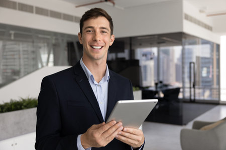 Young 30s businessman in professional attire standing in contemporary office, holding tablet,...
