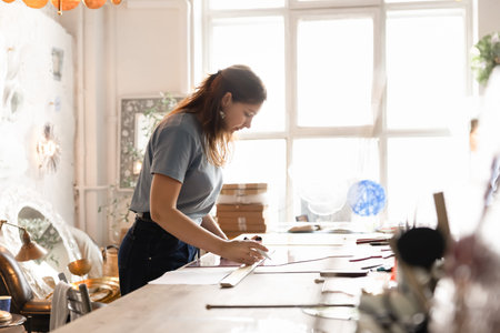 Female artisan working in creative workshop studio, sketching design on glass sheet, organizing...