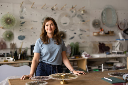 Smiling female artisan standing at workshop table, surrounded by handmade sconces, glasswork...
