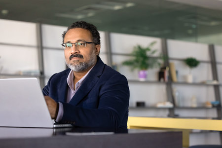 Focused middle-age businessman sits at desk with laptop during workday, thinking on information,...