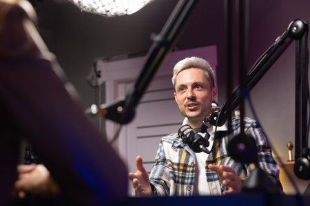 Male podcast host speaking into microphone while talking with guest during live recording session in...
